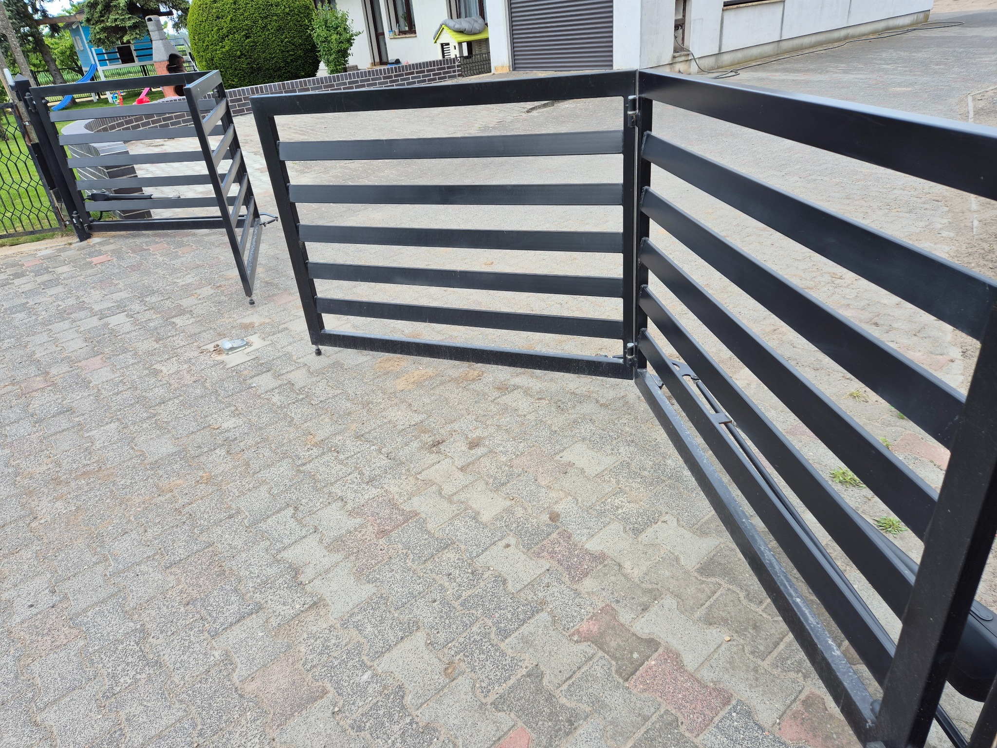 Nowoczesna brama wjazdowa, czarna, poziome profile, częściowo otwarta. Widoczny fragment posesji i kostka brukowa na podjeździe. Brama dwuskrzydłowa.