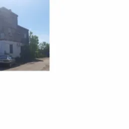 Dwukondygnacyjny budynek z okrągłą wieżą, obok zaparkowane samochody. Widoczne elementy infrastruktury energetycznej w tle. Jasne niebo, dzień.