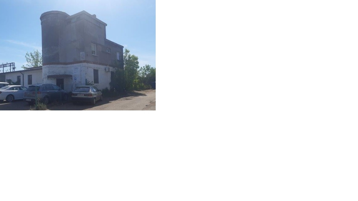 Dwukondygnacyjny budynek z okrągłą wieżą, obok zaparkowane samochody. Widoczne elementy infrastruktury energetycznej w tle. Jasne niebo, dzień.