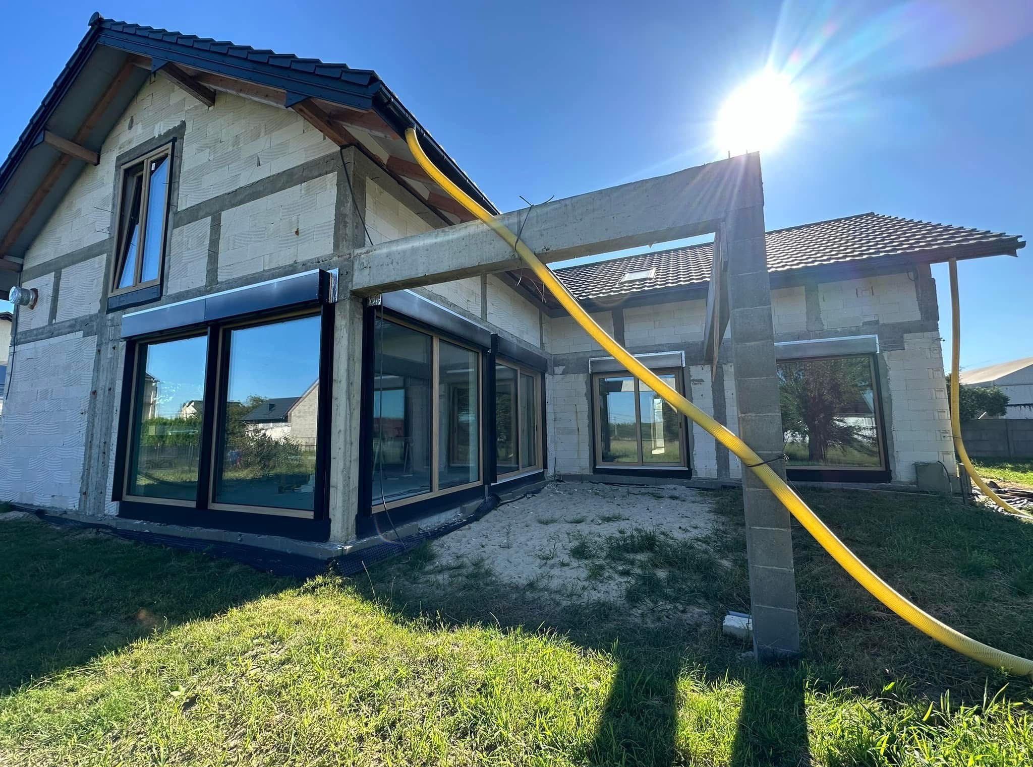 Dom w stanie surowym otwartym z zamontowanymi oknami z czarnymi ramami, na zewnątrz trawa i piasek, w tle błękitne niebo i słońce.