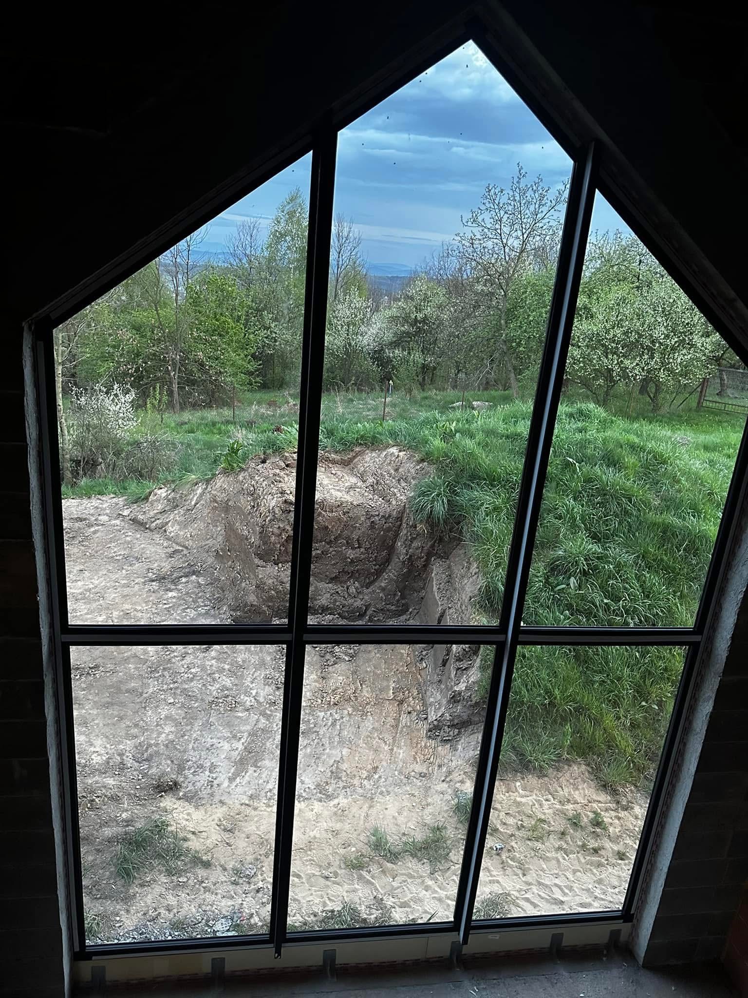 Trapezoidalne okno z czarną ramą, widok na ogród z ziemnym nasypem. Nietypowy kształt ramy i widok na zewnątrz.