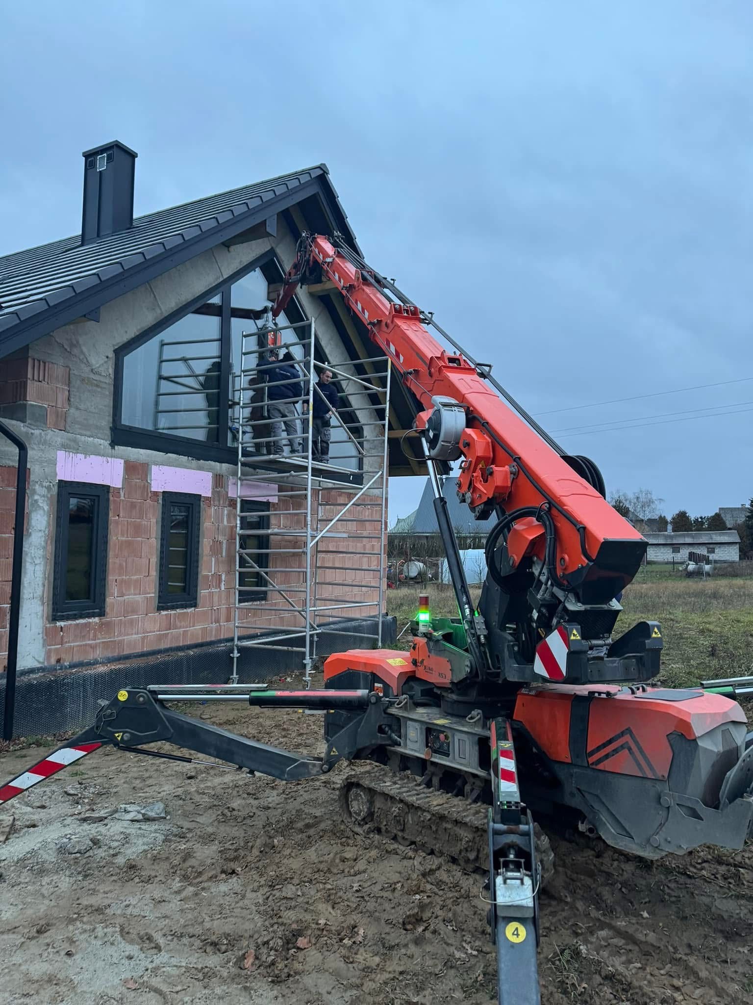 Montaż okna na surowym budynku z użyciem dźwigu gąsienicowego. Dwóch monterów na rusztowaniu. Widoczne okna z czarną ramą i elementy ocieplenia.