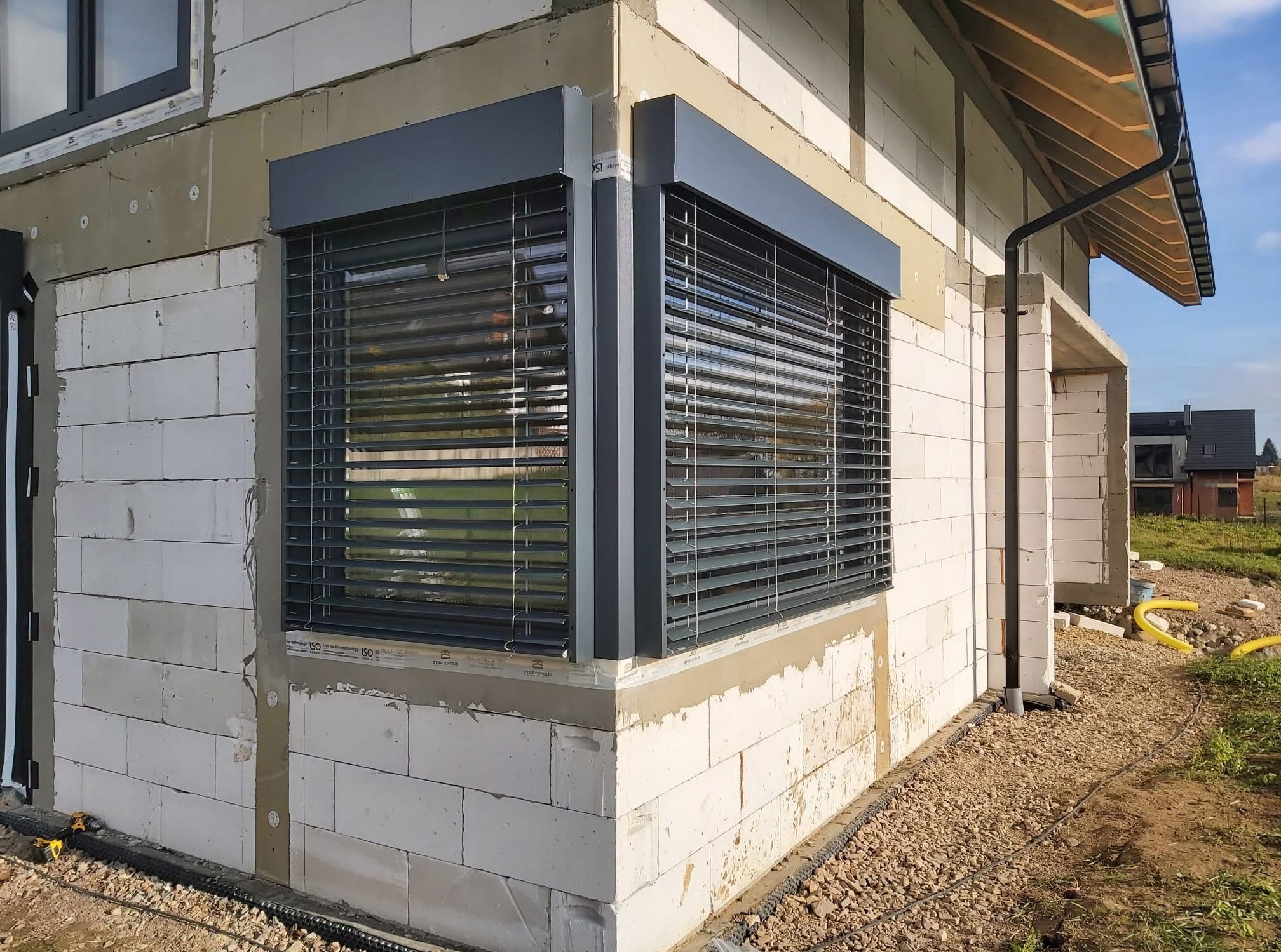 Narożne okno z szarymi roletami zewnętrznymi w surowym stanie budynku z białego bloczka. Widoczne okno PCV na górze. Na zewnątrz rozsypany gruz.