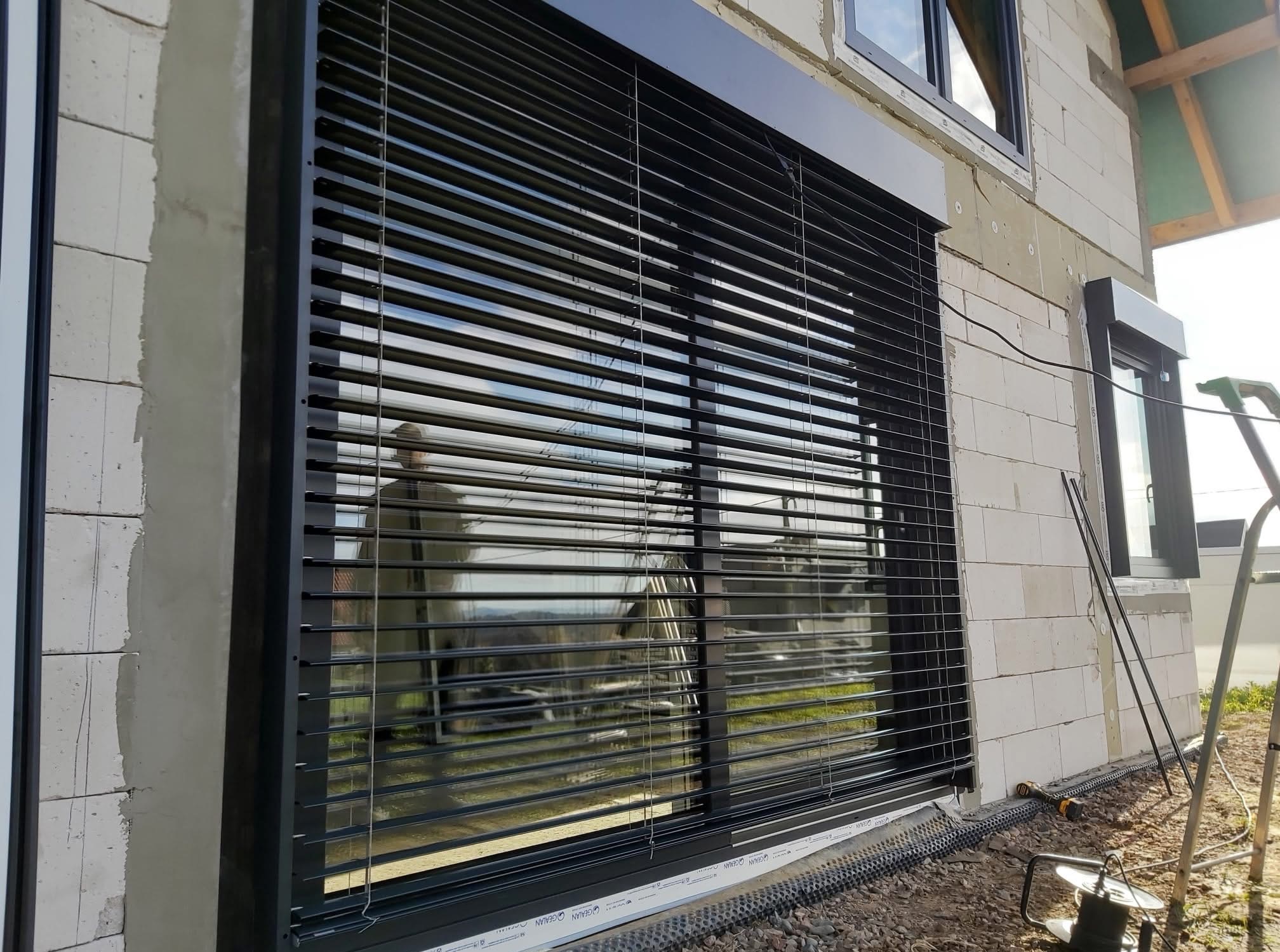 Czarne okno z żaluzjami fasadowymi w surowym budynku z białego gazobetonu. Widoczne narzędzia i drabina. Okno z roletą zewnętrzną.