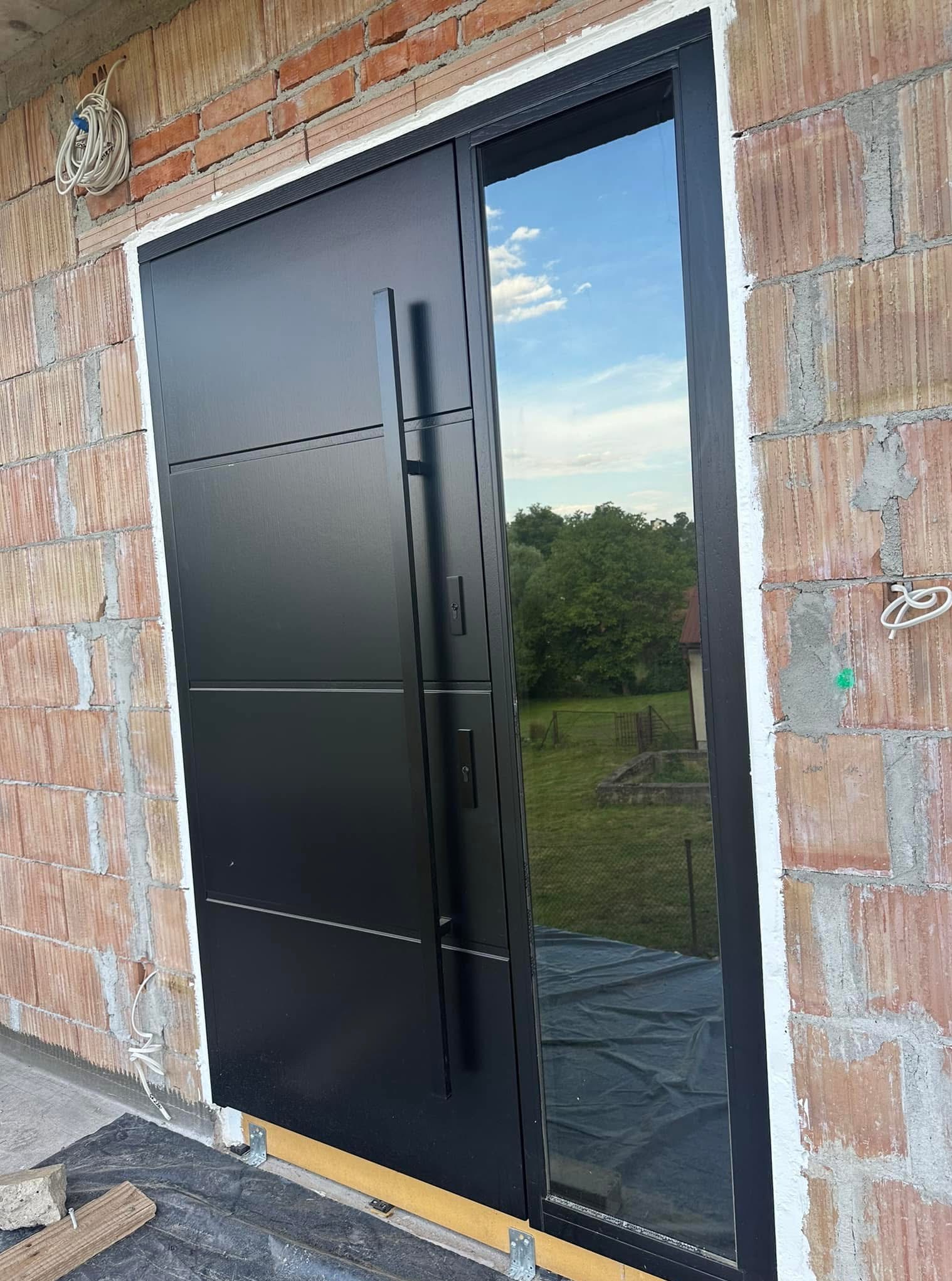 Czarne drzwi zewnętrzne z długim uchwytem i szybą, zamontowane w surowej ścianie z cegły, odbijające widok nieba i zieleni. Nowoczesny design.