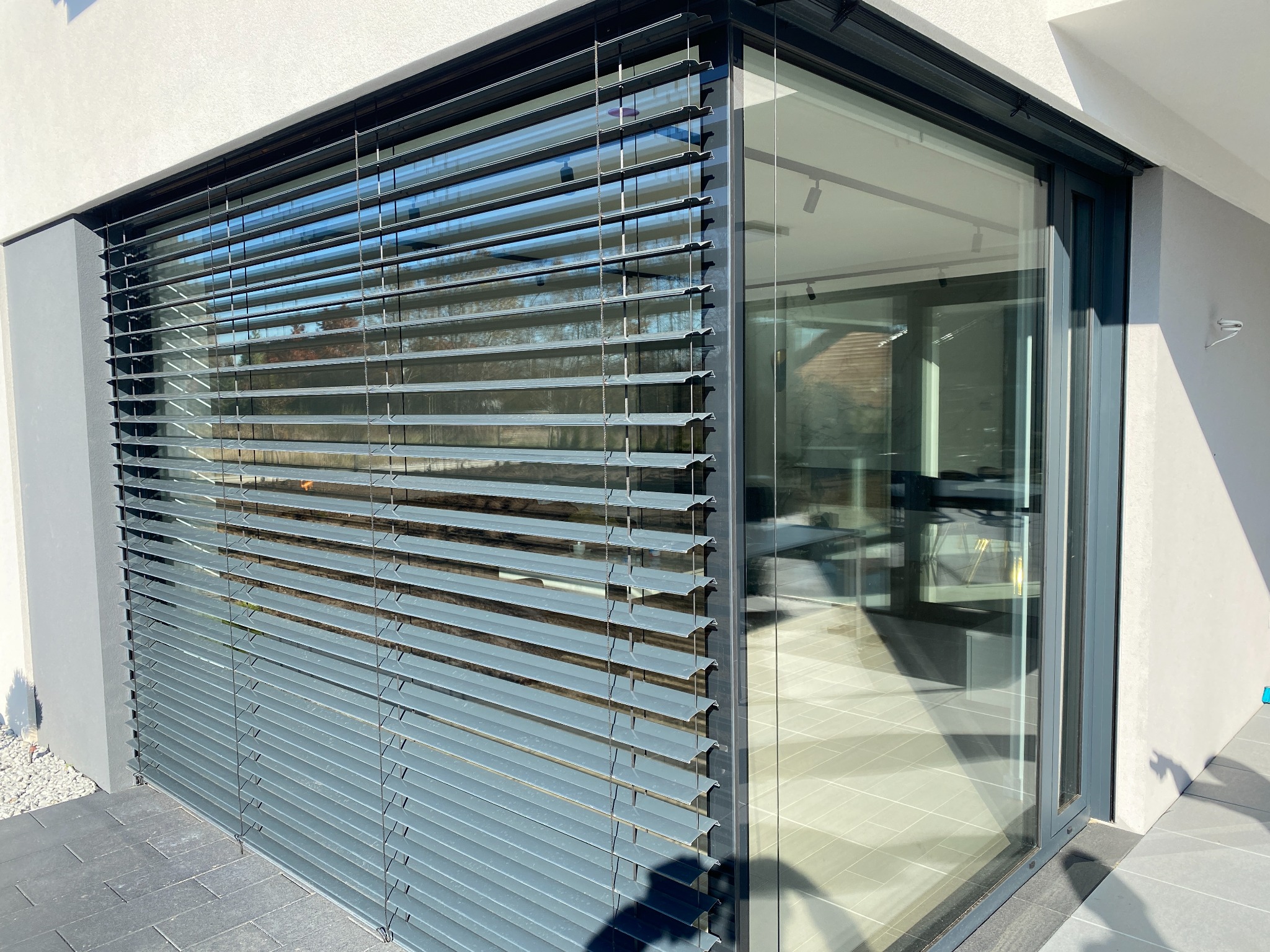 Nowoczesne, szare żaluzje poziome zamontowane na dużym oknie narożnym, z widokiem na wnętrze domu. Minimalistyczny design, jasne światło dzienne.