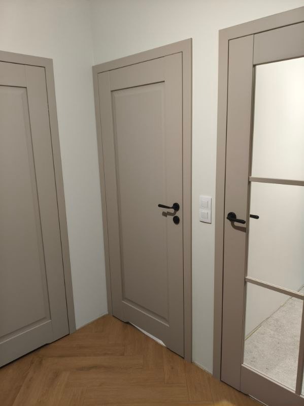 Trzy beżowe drzwi wewnętrzne z czarnymi klamkami w korytarzu z parkietem ułożonym w jodełkę. Jedne drzwi przeszklone z podziałami.