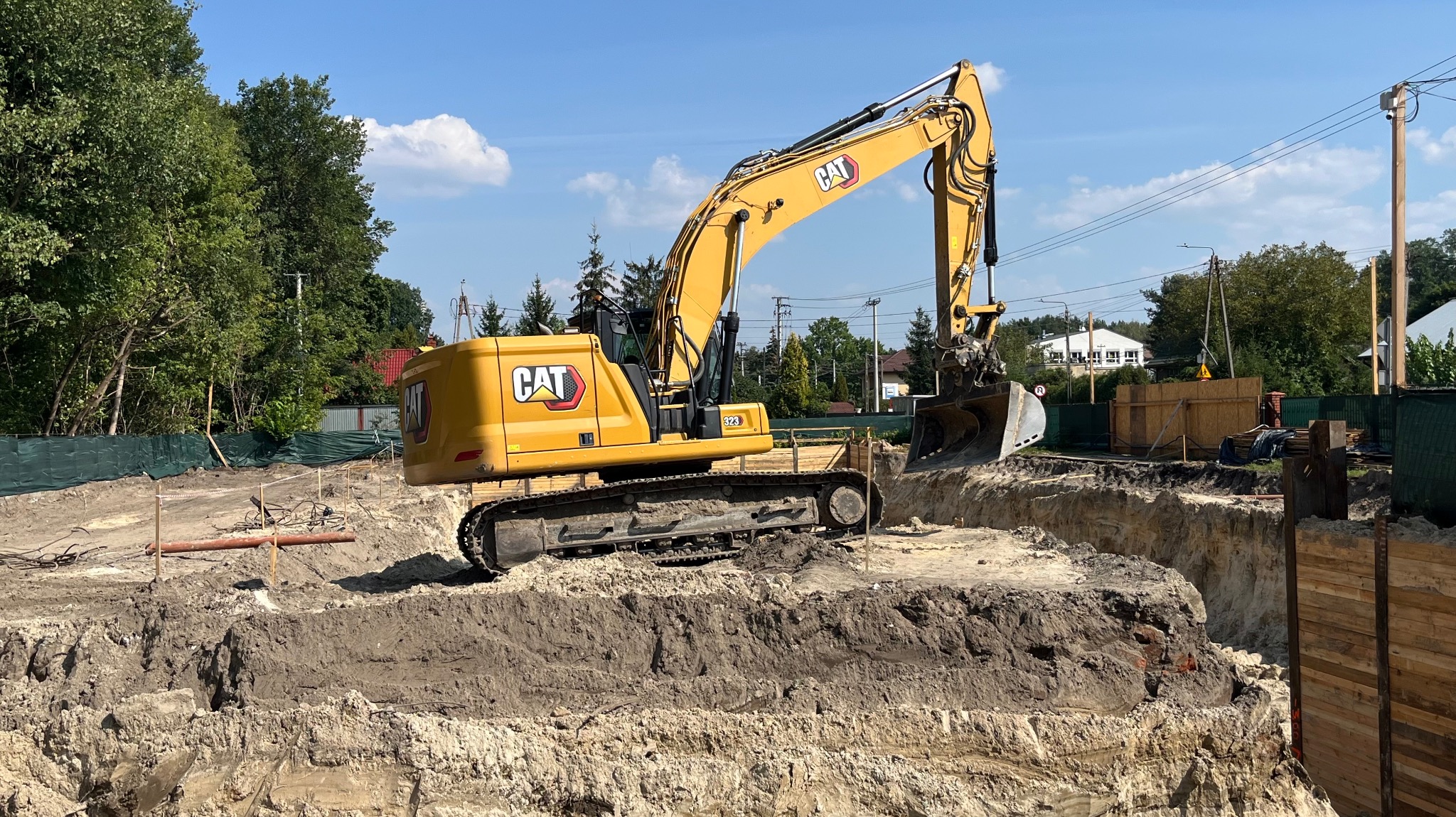 Żółta koparka CAT 323 na placu budowy, wykonująca wykop pod fundamenty. Widoczne przygotowane zabezpieczenia wykopu i elementy infrastruktury.