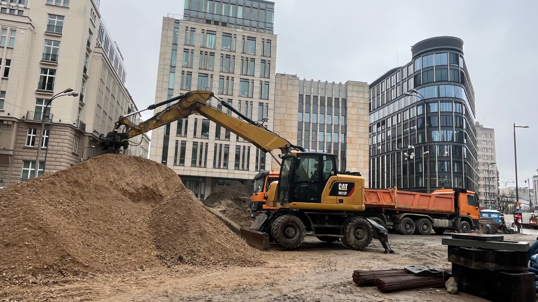 Żółta koparka CAT M315F z pomarańczową wywrotką na tle miejskiej zabudowy. Górka piachu i stalowe pręty na pierwszym planie.