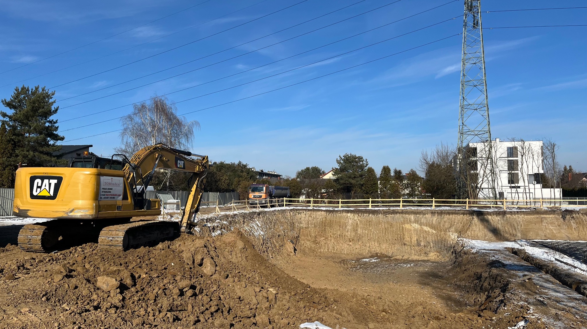 Żółta koparka CAT na skraju wykopu pod fundamenty, z widocznym transportem ziemi ciężarówką i budynkiem w tle, w słoneczny dzień.