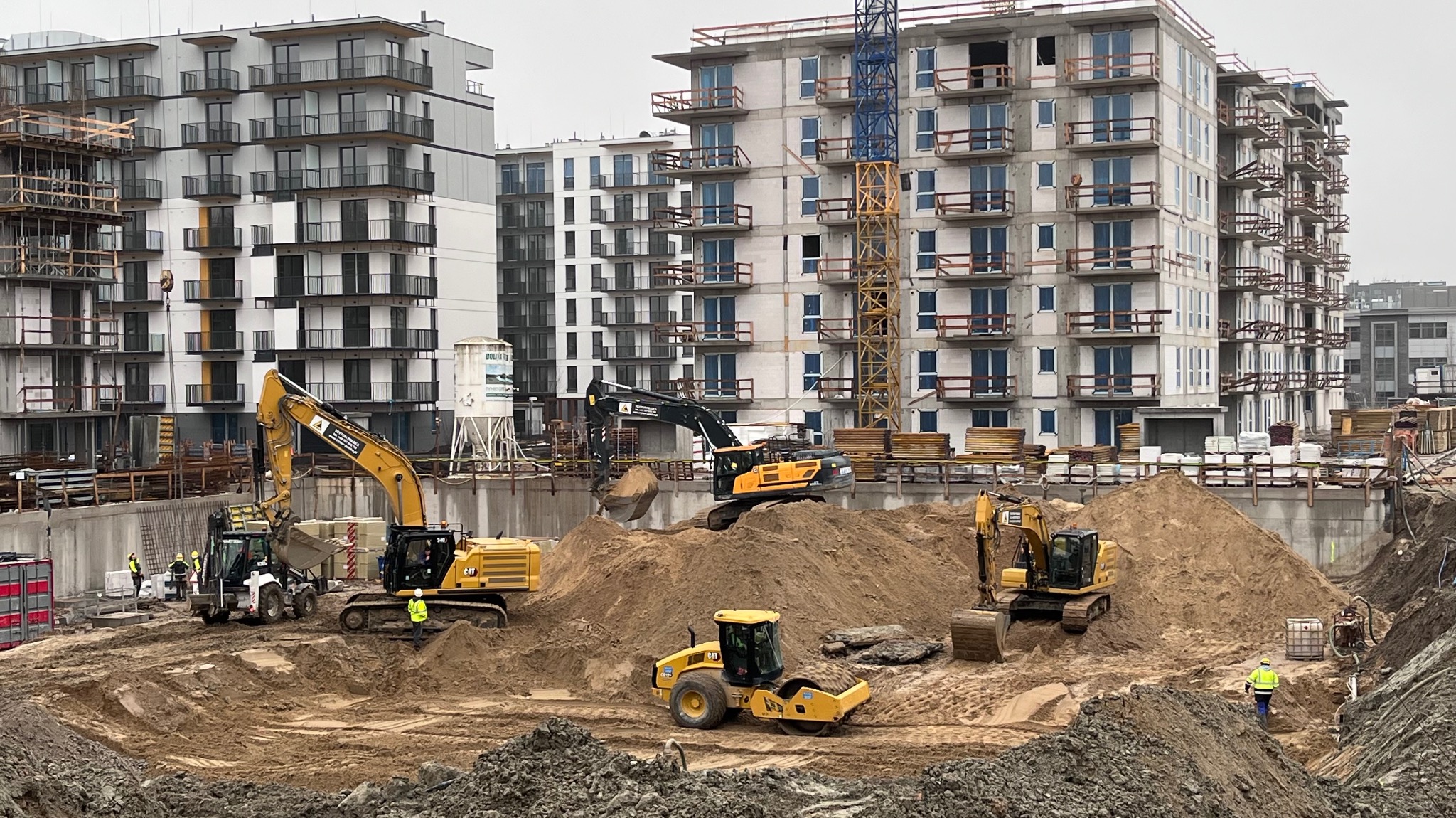 Budowa osiedla mieszkaniowego: praca kilku koparek i walca na rozległym placu budowy, na tle wznoszonych budynków mieszkalnych w pochmurny dzień.