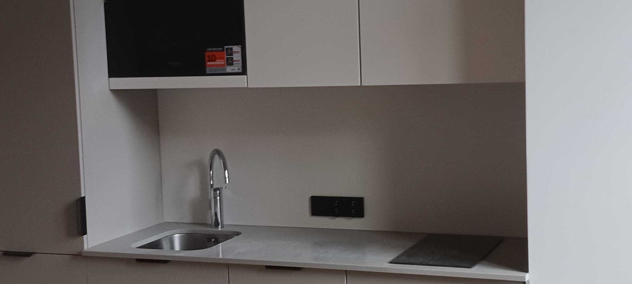 Nowoczesna, minimalistyczna kuchnia z białymi szafkami, chromowaną baterią i czarną mikrofalówką. Jasny blat i subtelne oświetlenie podkreślają czystość designu.