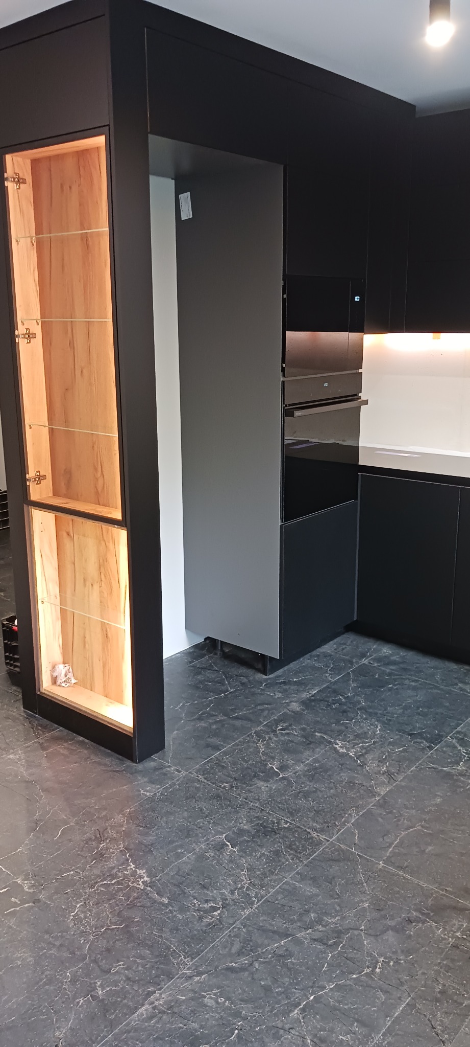 Nowoczesna, czarna zabudowa kuchenna z drewnianą witryną oświetloną od wewnątrz. Na podłodze szare płytki imitujące marmur. Widoczny piekarnik w zabudowie.