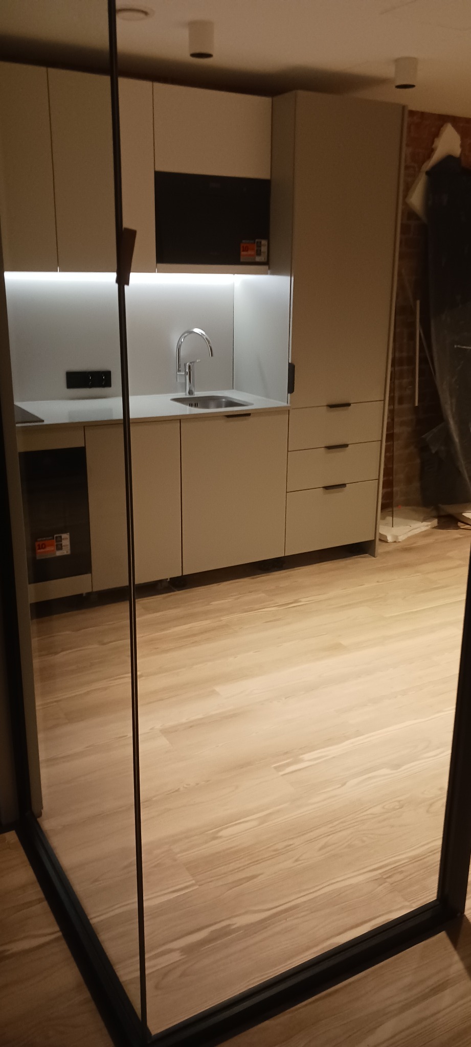 Jasna kuchnia z zabudową pod sufit, minimalistycznym zlewem i drewnianą podłogą, widziana przez szklane drzwi. Oświetlenie LED podkreśla czystość linii.