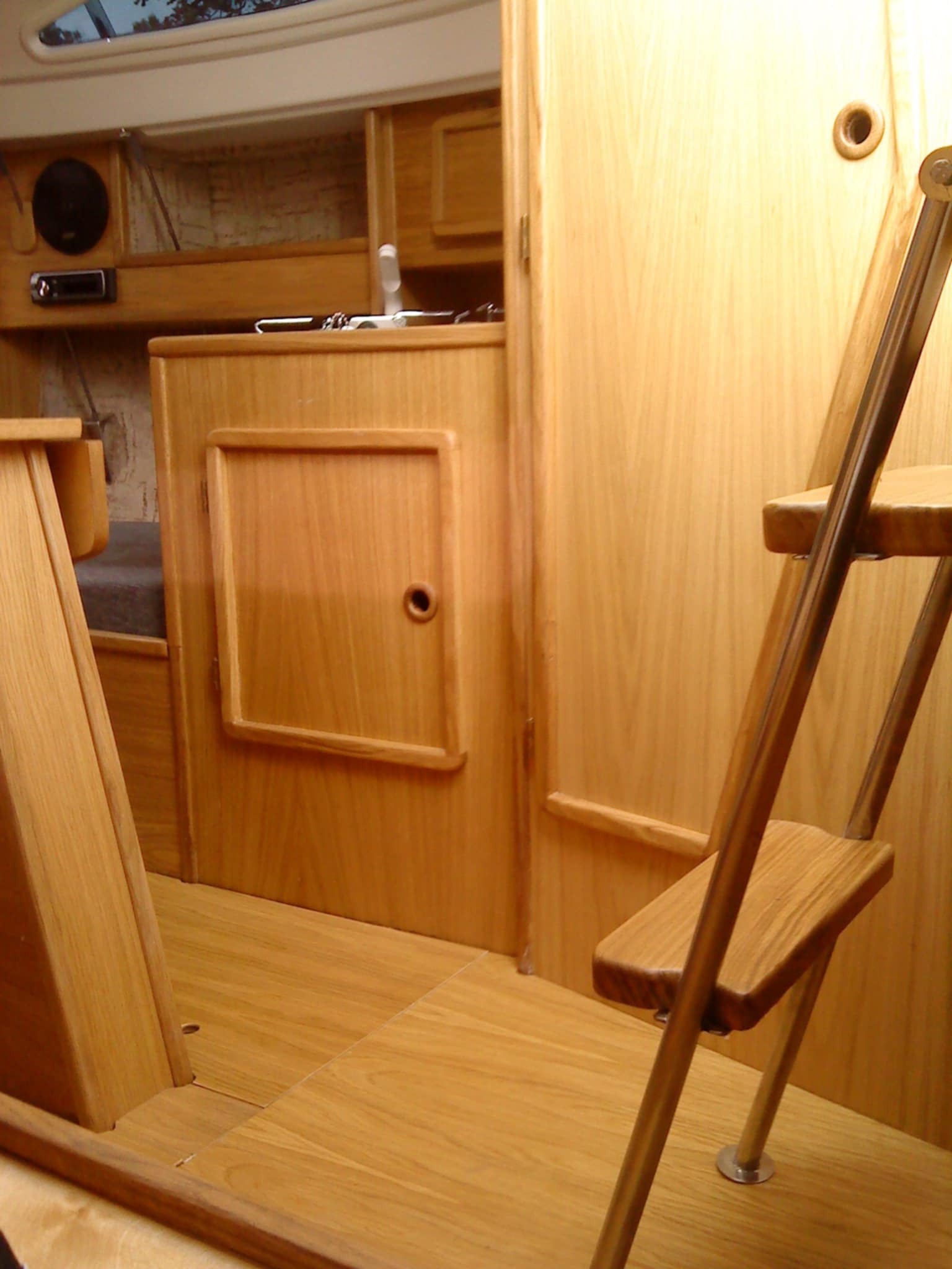 Drewniane wnętrze małej kabiny z drabinką, szafką z okrągłym otworem zamiast uchwytu i radiem. Światło wpada przez okno dachowe.