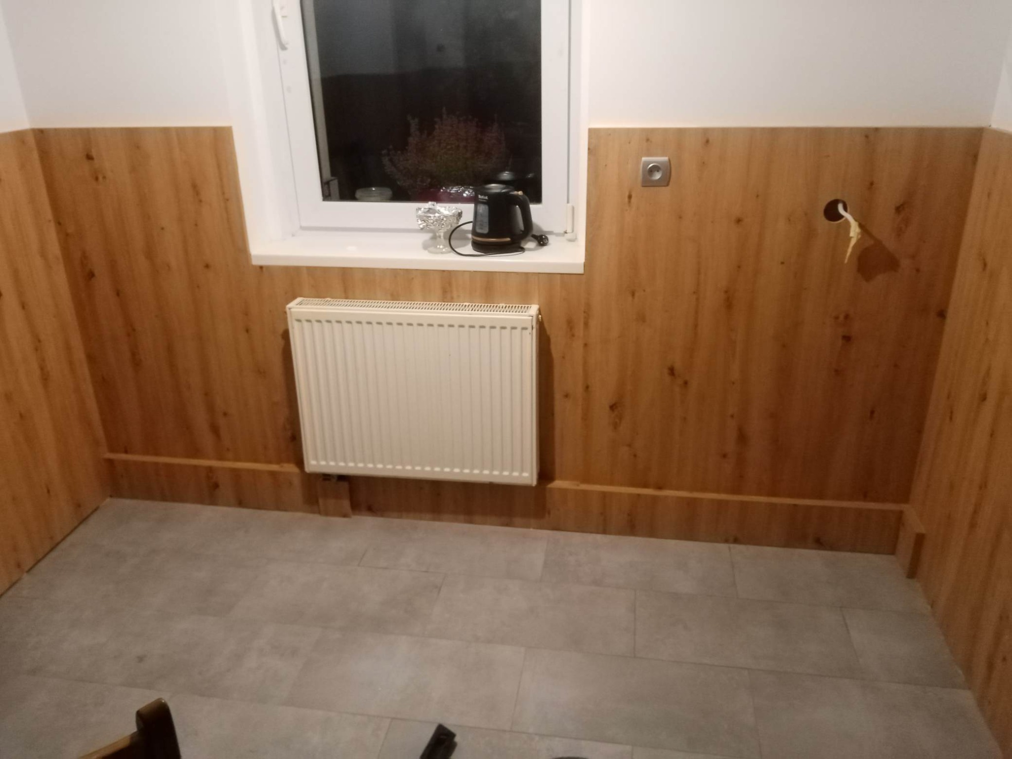 Wykończone panele ścienne z efektem drewna w pomieszczeniu z grzejnikiem i szarymi płytkami podłogowymi. Widoczne gniazdko i otwór na kabel w ścianie.