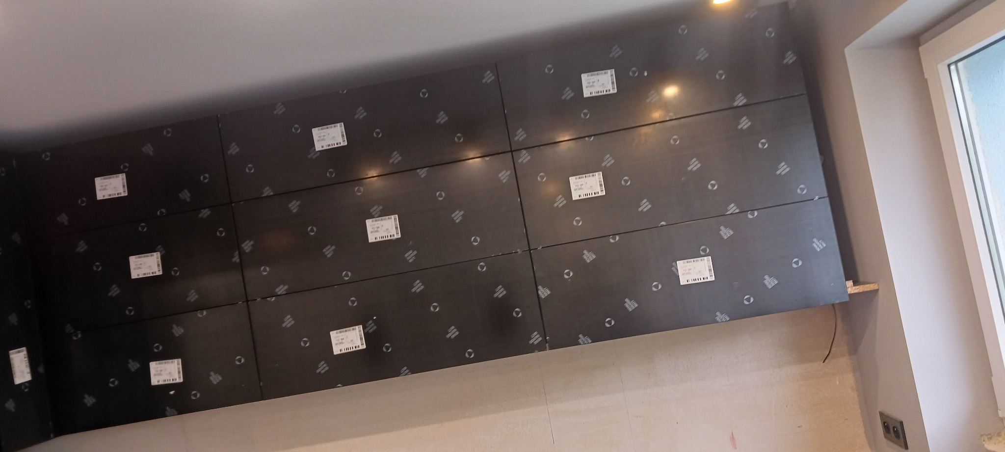 Montaż czarnych paneli ściennych z naklejkami w jasnym wnętrzu, widoczne okno i gniazdko elektryczne. Etap prac wykończeniowych, możliwe ocieplenie lub przygotowanie pod montaż mebli.