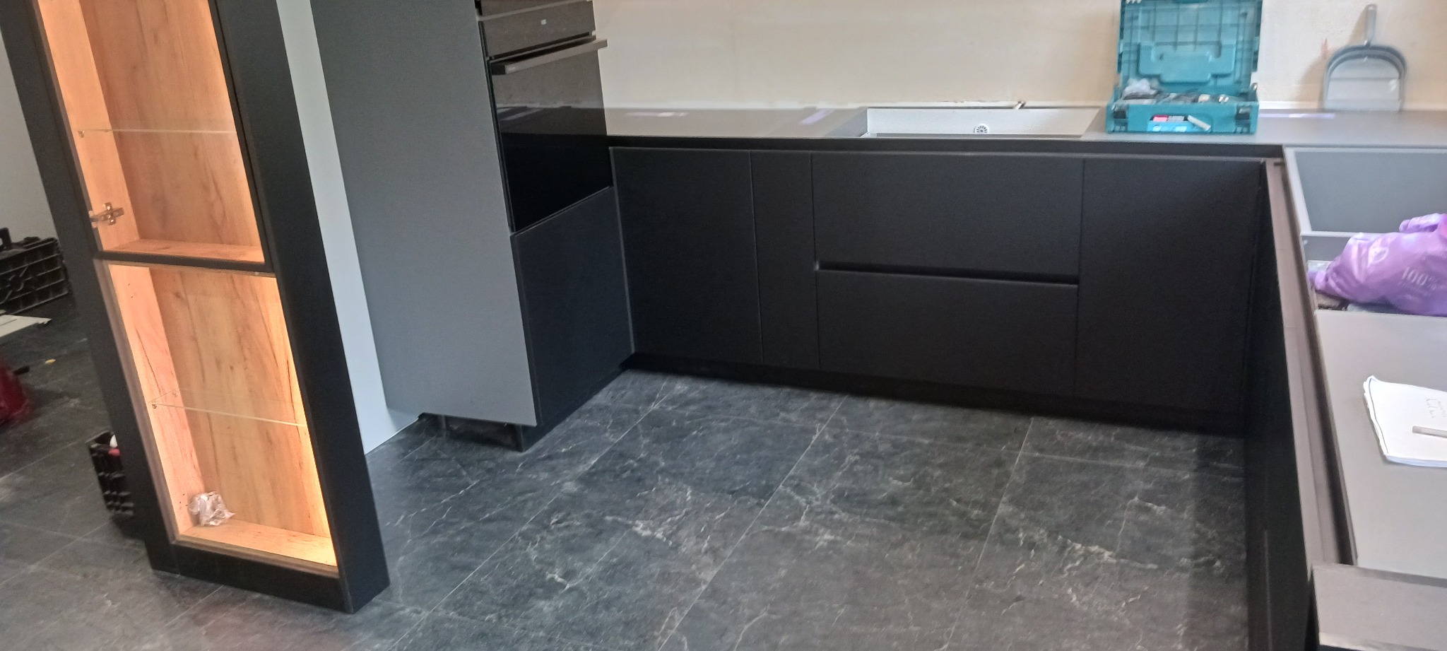 Częściowy widok nowoczesnej, czarnej kuchni z podświetlaną witryną i szarymi płytkami podłogowymi imitującymi marmur. Na blacie skrzynka z narzędziami.