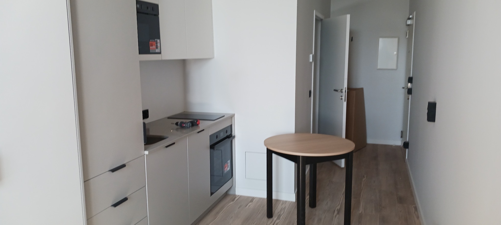 Jasna kuchnia z szarymi frontami, okrągły stół z czarnymi nogami. Na blacie wiertarka. W tle otwarte drzwi i karton. Minimalistyczne wykończenie wnętrza.