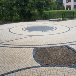 Układanka z kostki granitowej w beżach i szarościach tworzy dekoracyjny wzór na podjeździe. Okrągły motyw centralny i obrzeża z ciemnej kostki.