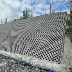 Skarpa wzmocniona ażurowymi płytami betonowymi na wzgórzu, zabezpieczona siatką ogrodzeniową. Niebieski przewód u podnóża. Widok na pochmurne niebo.