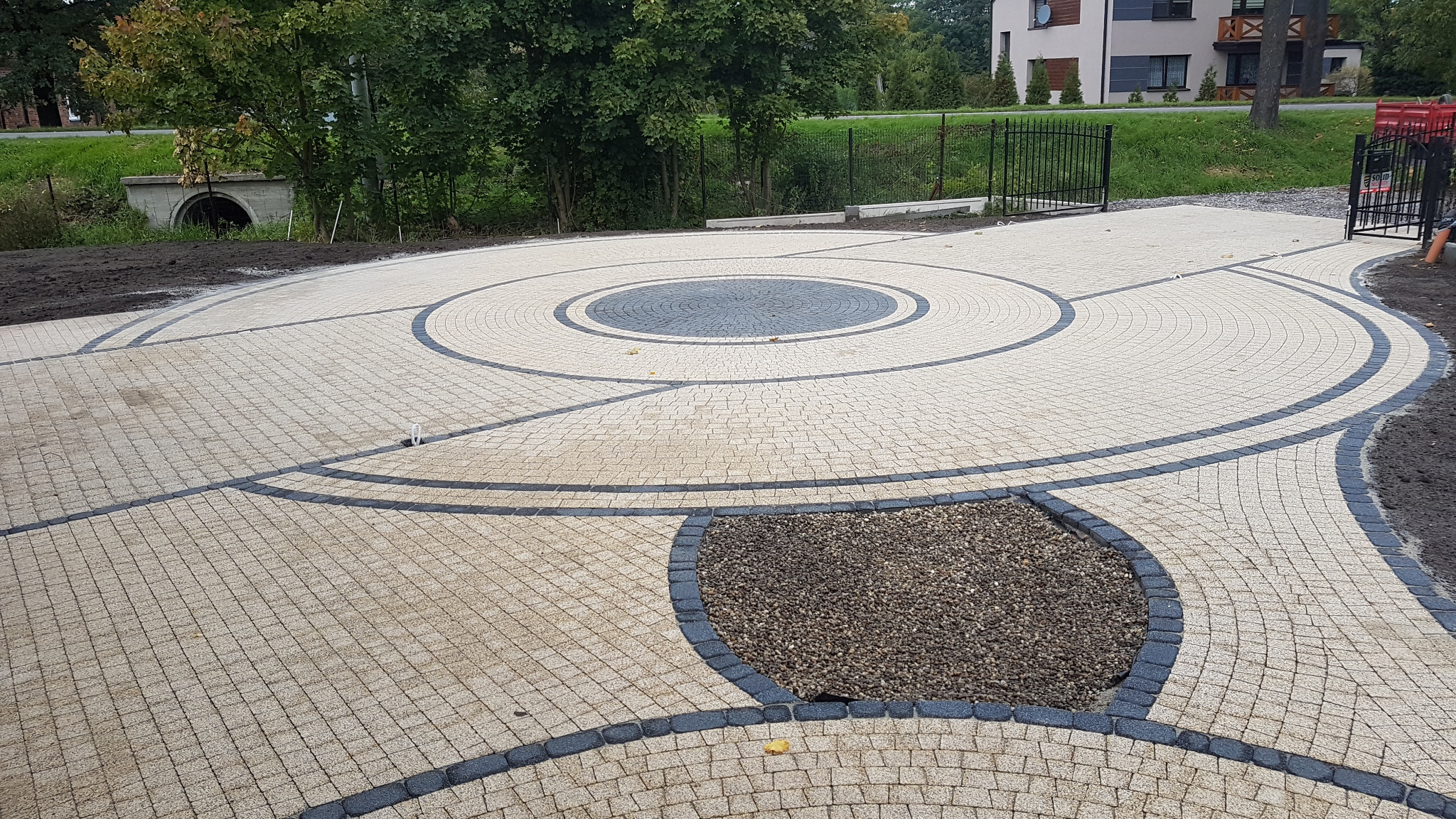 Układanka z kostki granitowej w beżach i szarościach tworzy dekoracyjny wzór na podjeździe. Okrągły motyw centralny i obrzeża z ciemnej kostki.