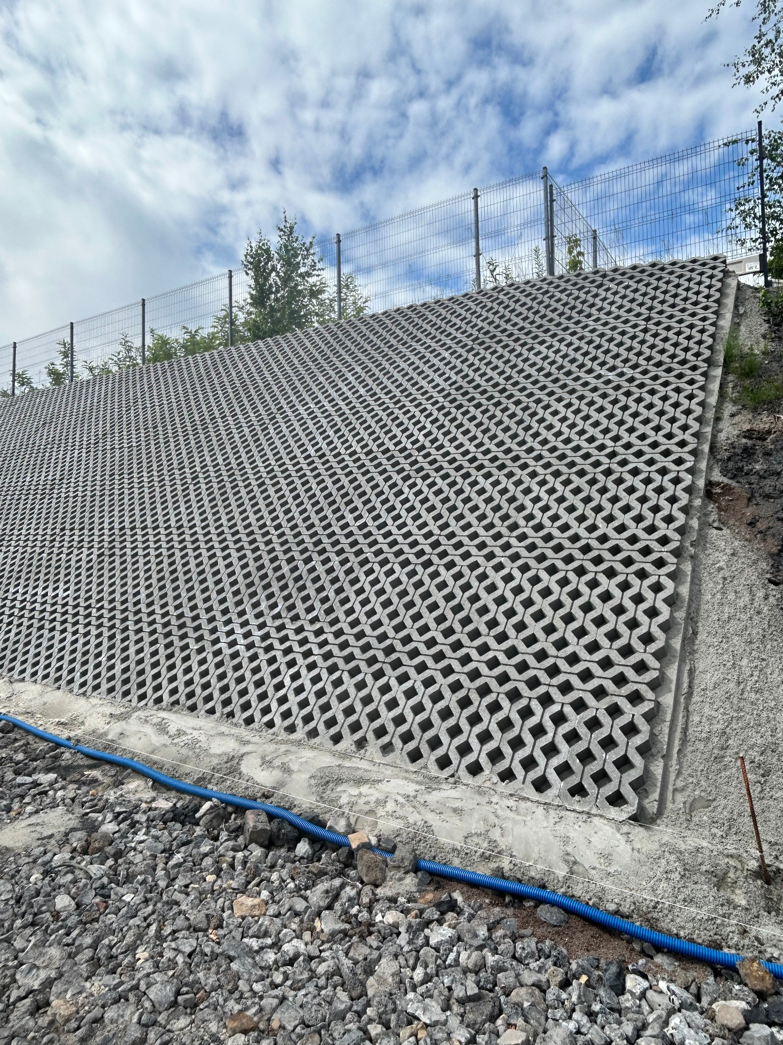 Skarpa wzmocniona ażurowymi płytami betonowymi na wzgórzu, zabezpieczona siatką ogrodzeniową. Niebieski przewód u podnóża. Widok na pochmurne niebo.