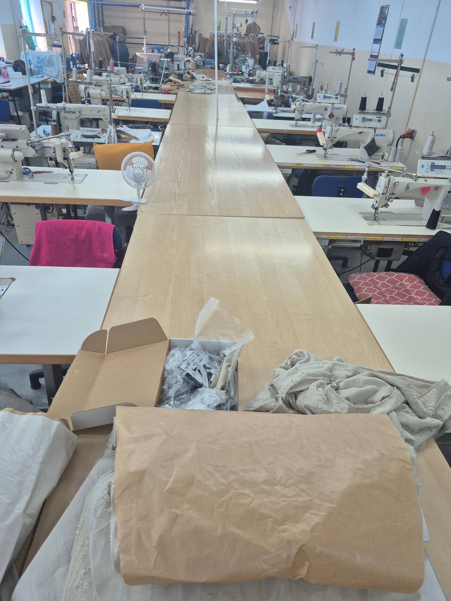 Szwalnia: Długi stół krojczy z materiałami i akcesoriami. W tle liczne maszyny do szycia. Widok na przestronną halę produkcyjną.