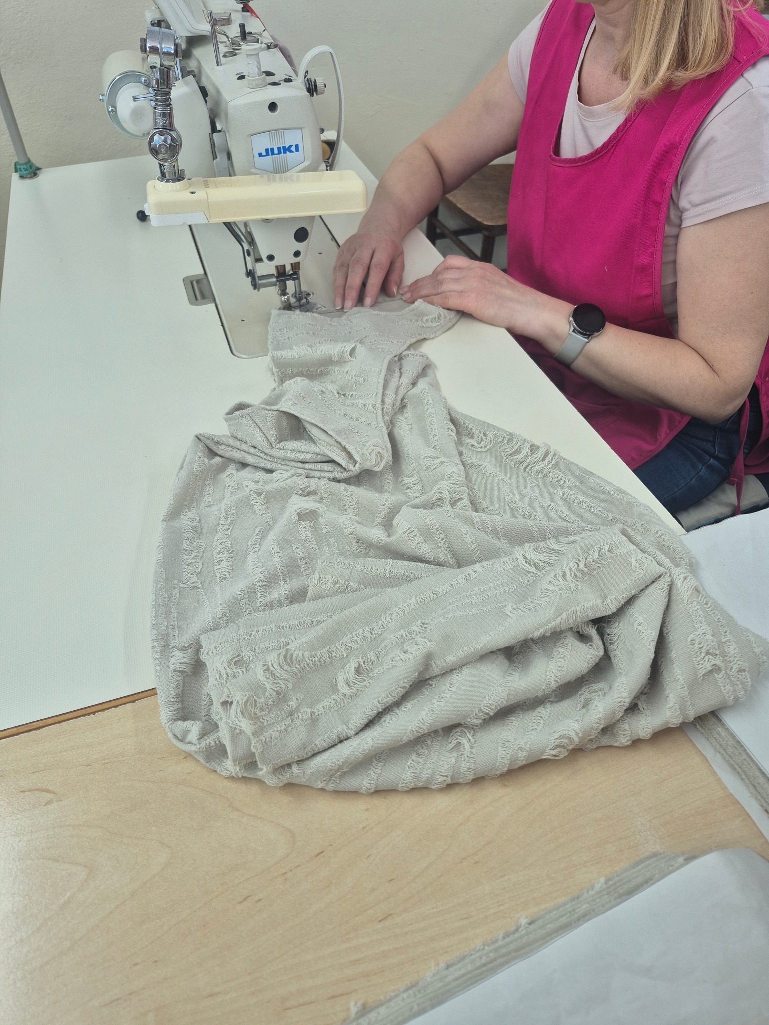Praca przy maszynie JUKI. Osoba w różowym fartuchu szyje jasną tkaninę z frędzlami. Widoczny fragment stołu i maszyny.