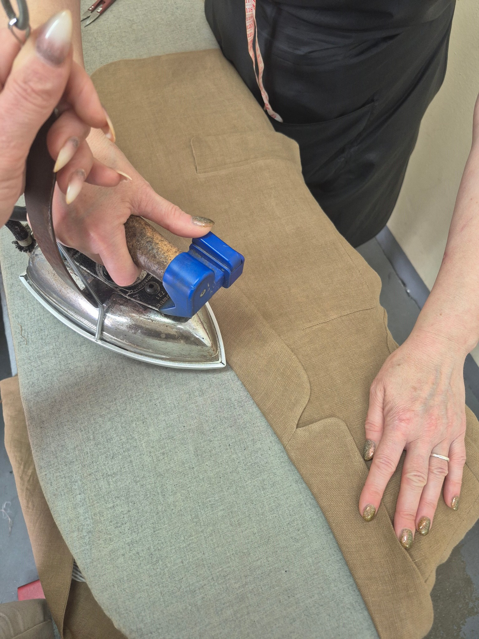 Prasowanie brązowych spodni z kieszenią na szarej desce staromodnym żelazkiem. Widoczne dłonie z długimi paznokciami i pierścionkiem.