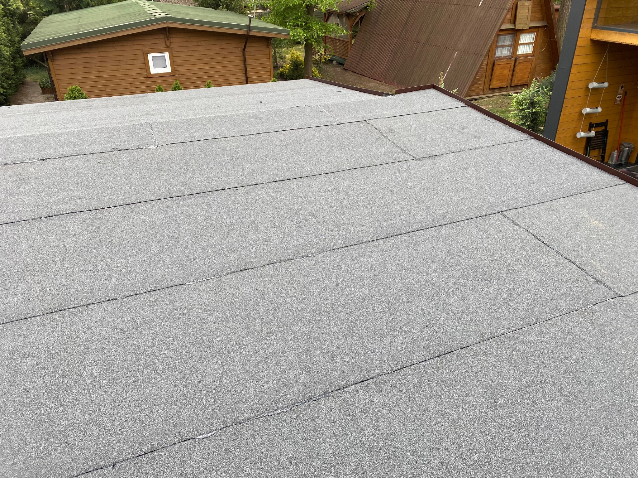 Szary dach płaski pokryty papą termozgrzewalną, z widocznymi łączeniami, na tle zabudowań i zieleni. Widok z góry, oświetlenie dzienne.