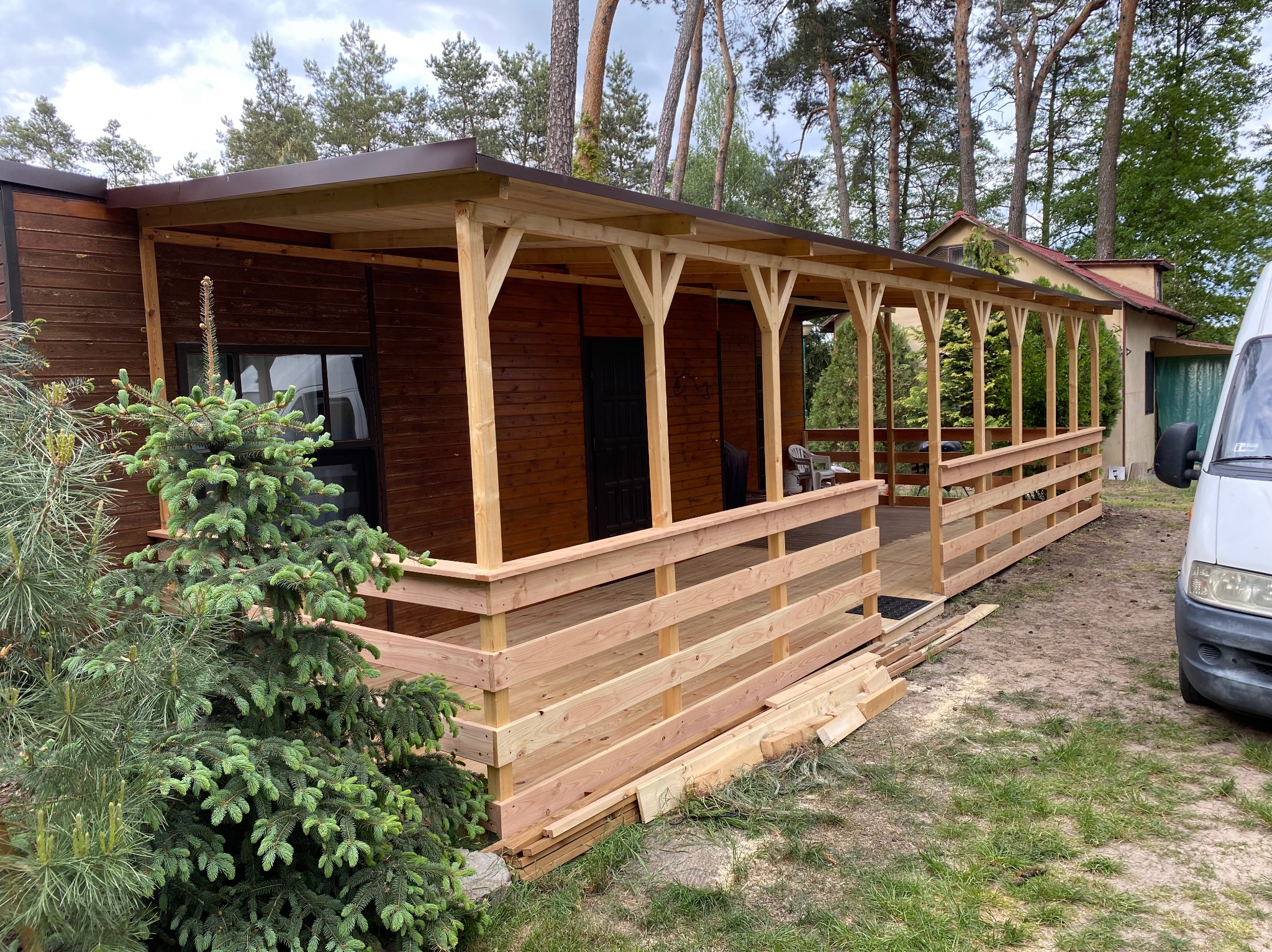 Drewniana altana z zadaszeniem przy brązowym domu, otoczona zielenią drzew i krzewów. Widoczne elementy konstrukcyjne w naturalnym kolorze drewna. Częściowo widoczny samochód dostawczy.