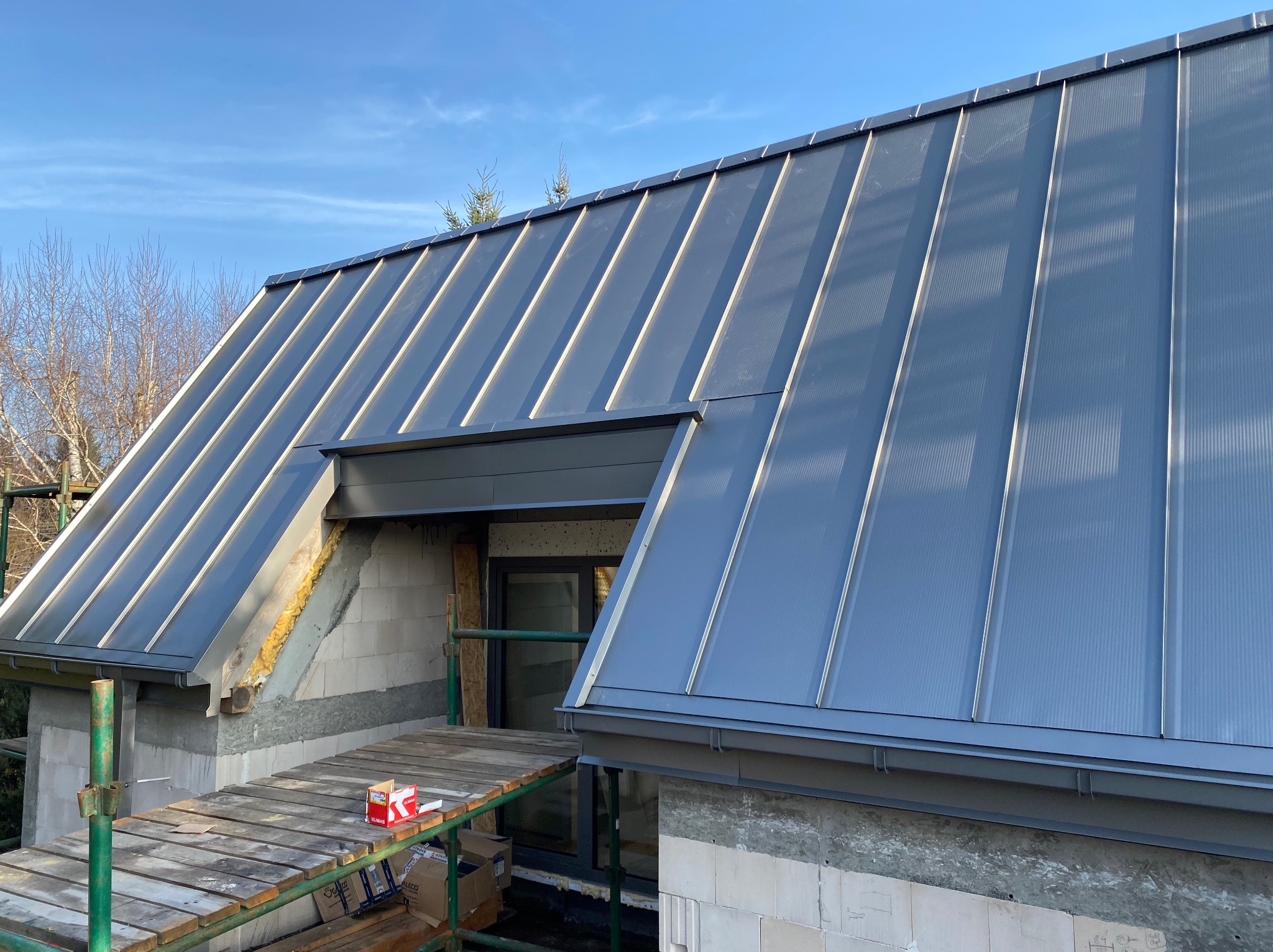 Szary dach z blachy trapezowej na budynku w trakcie budowy. Widoczne rusztowania i materiały budowlane. Słoneczny dzień, błękitne niebo.