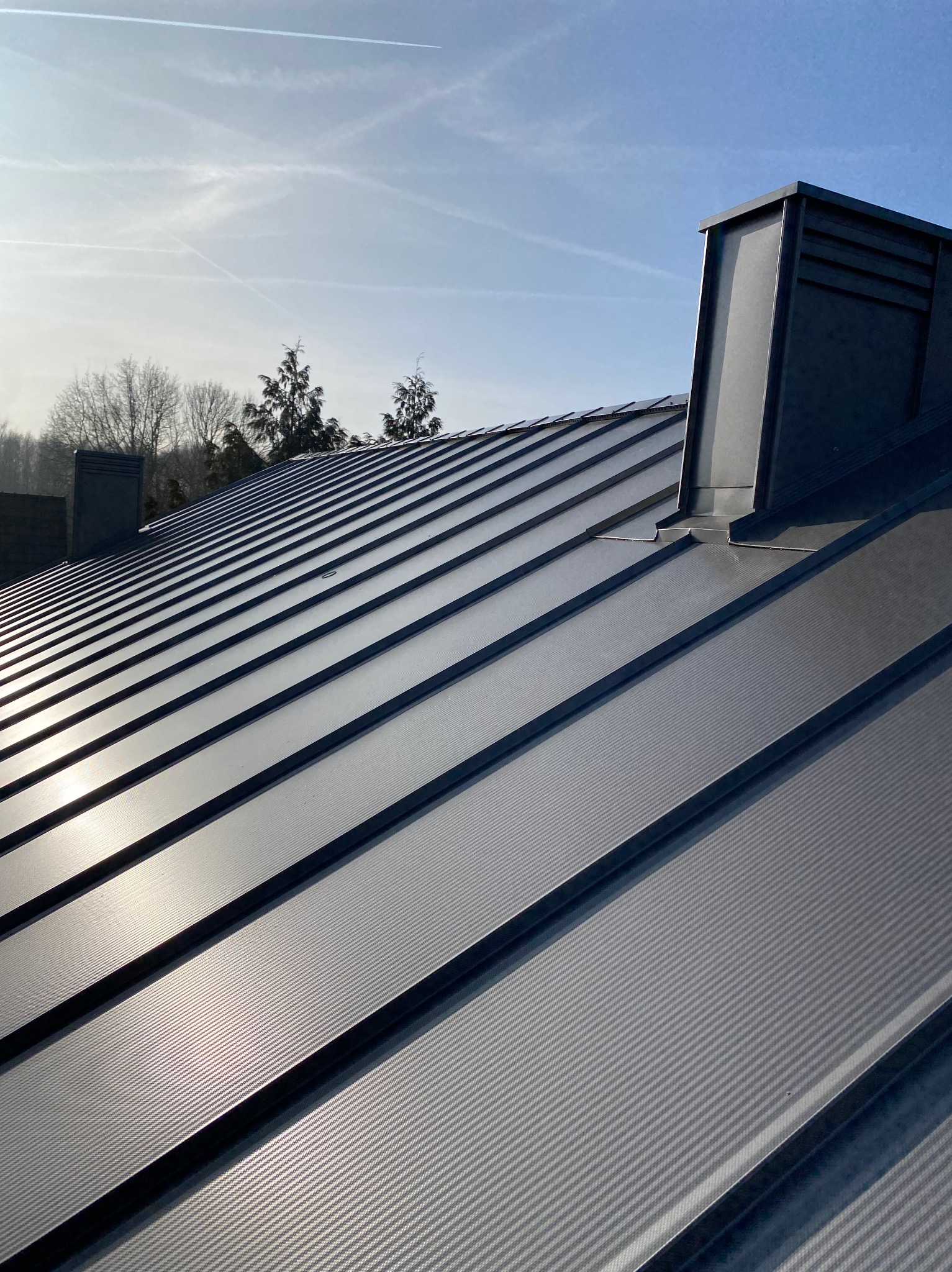 Szary, metalowy dach panelowy z kominem w słoneczny dzień. Widoczne drzewa w tle i smugi kondensacyjne na niebie. Nowoczesne pokrycie dachowe.