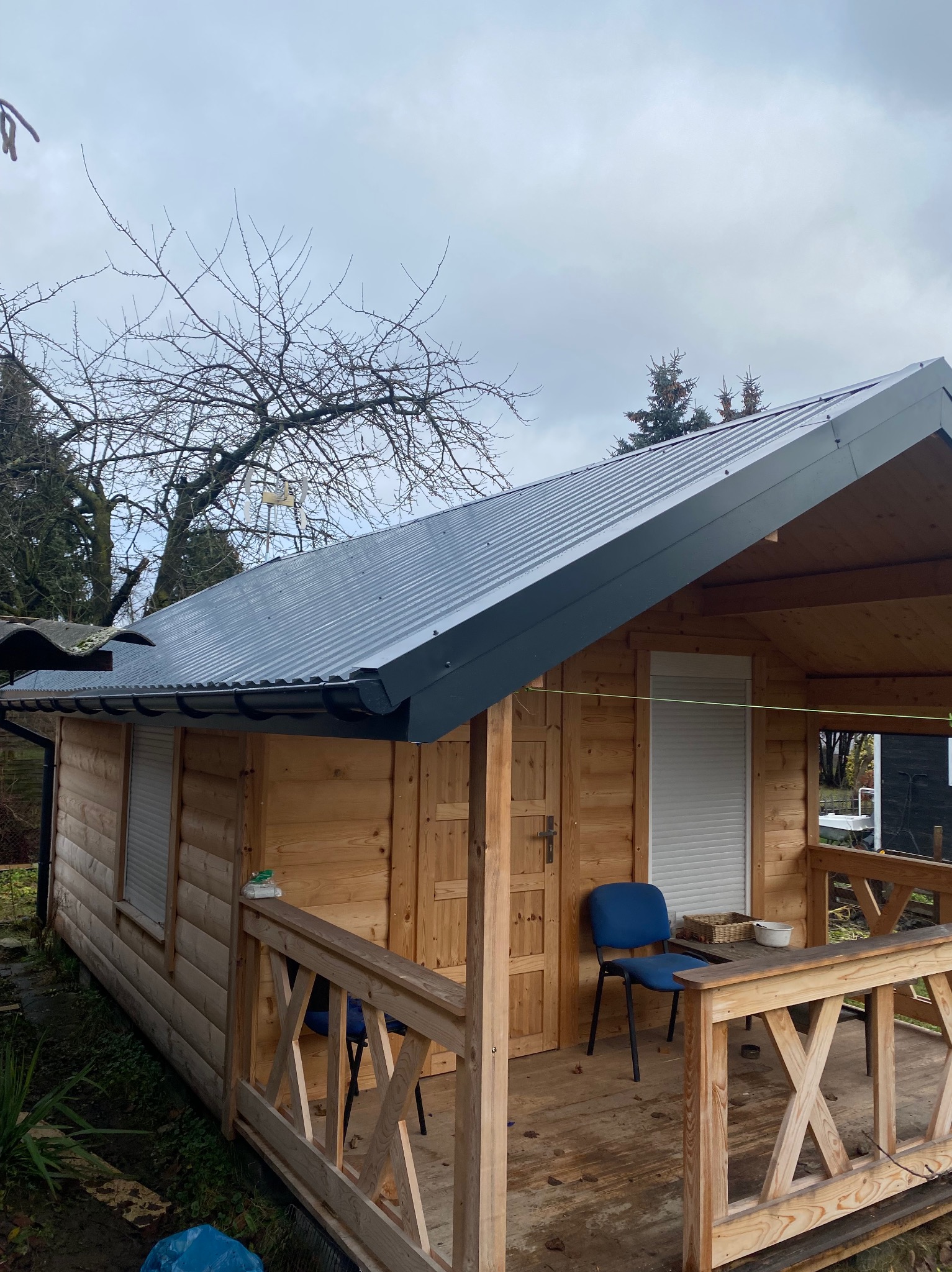 Drewniany domek z szarą, metalową dachówką. Widoczny zadaszony taras z drewnianą balustradą i niebieskim krzesłem. Tło stanowią drzewa i pochmurne niebo.