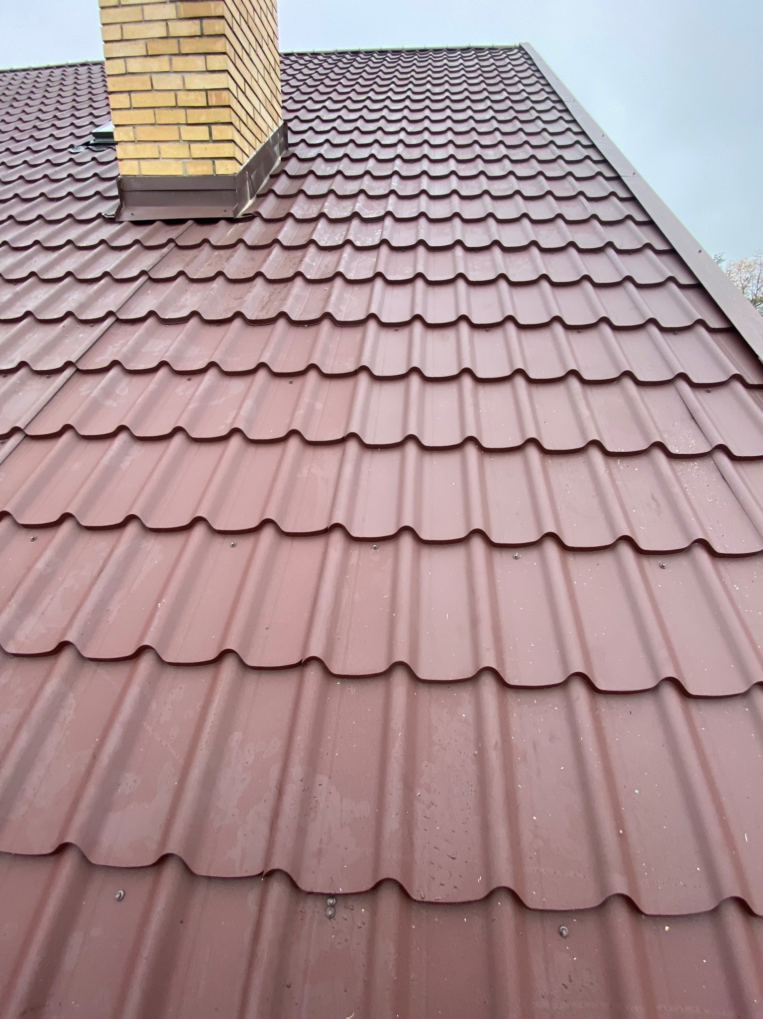 Czerwono-brązowy dach z blachy trapezowej, widok z dołu, z kominem z cegły klinkierowej. Widoczne mocowania blachy. Pochmurne niebo w tle.