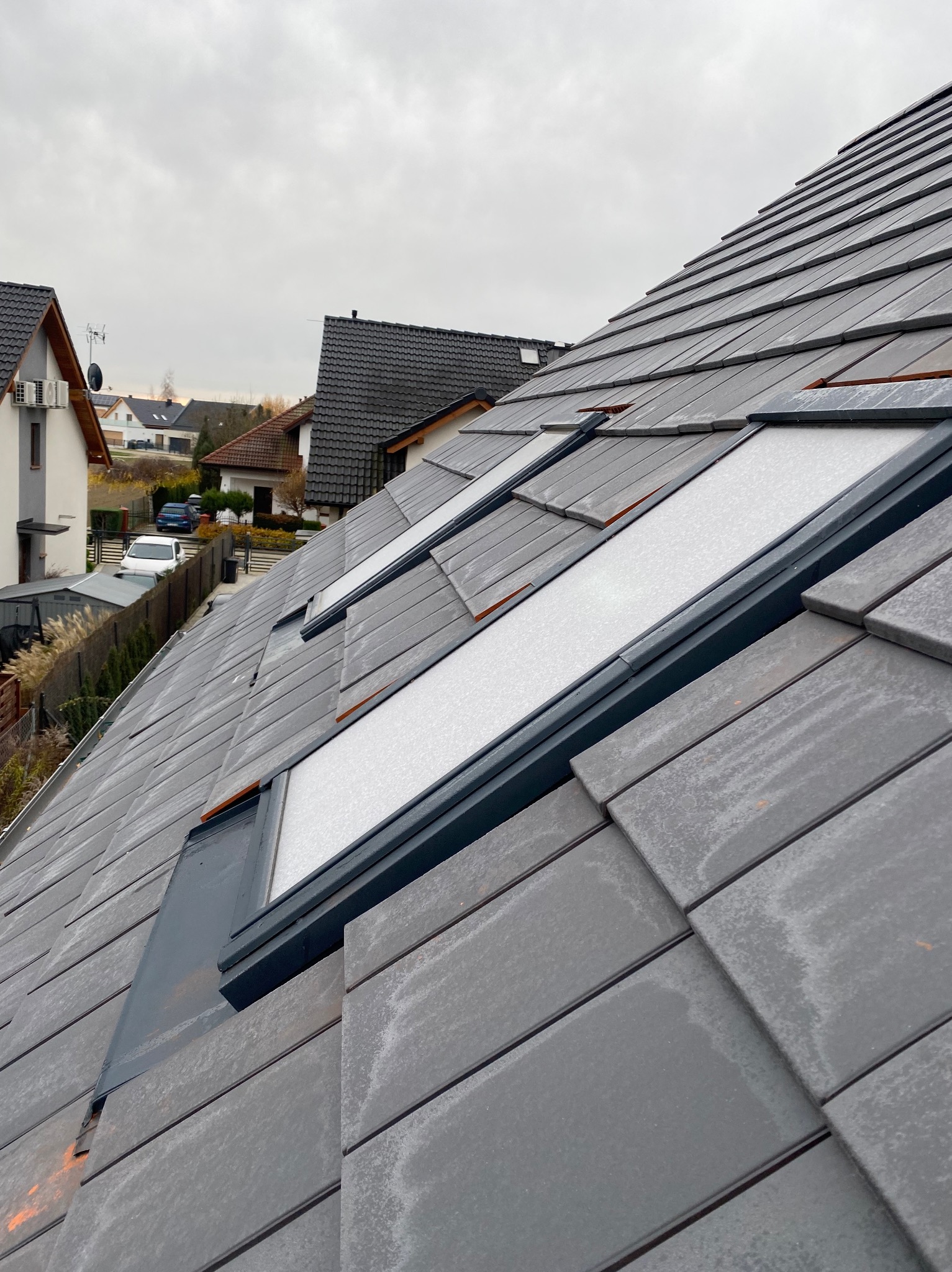 Szary dach z dachówką ceramiczną i trzema oknami dachowymi. Widok na domy w tle przy pochmurnej pogodzie. Dach lekko wilgotny, możliwe po deszczu.