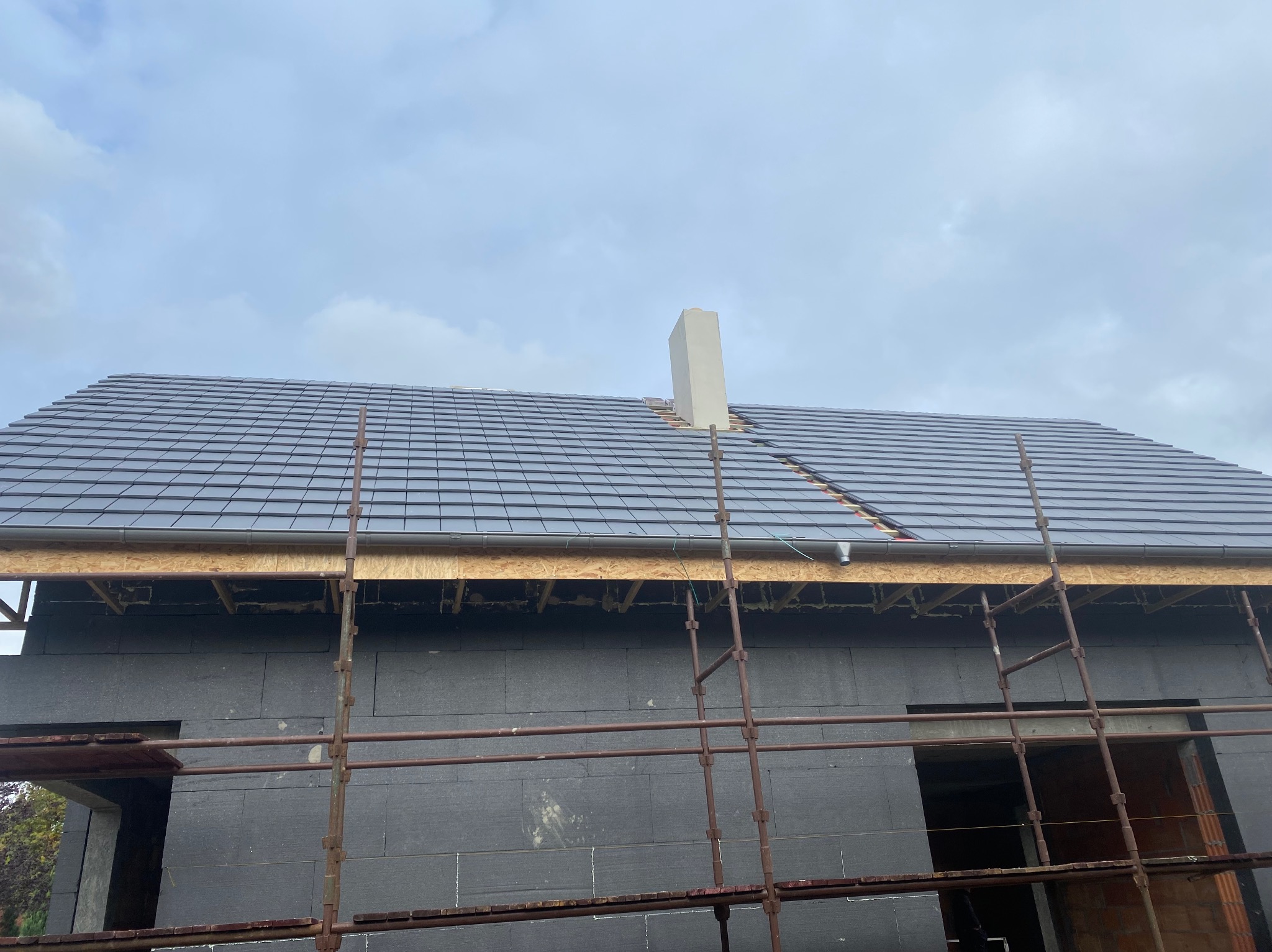 Nowy, grafitowy dach z kominem w trakcie budowy domu. Widoczne rusztowania i ocieplenie ścian. Ujęcie z dołu na tle pochmurnego nieba.