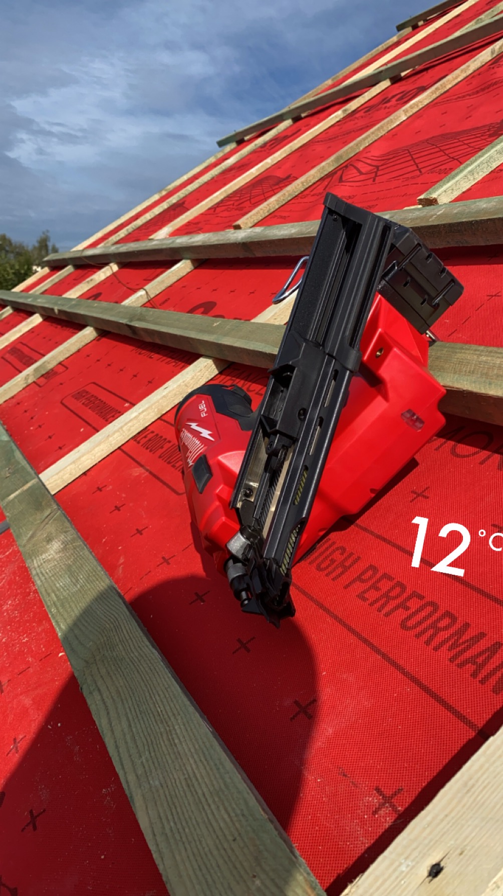 Czerwona membrana dachowa z listwami i narzędziem do wbijania gwoździ. Widoczne niebo i temperatura 12 stopni Celsjusza.