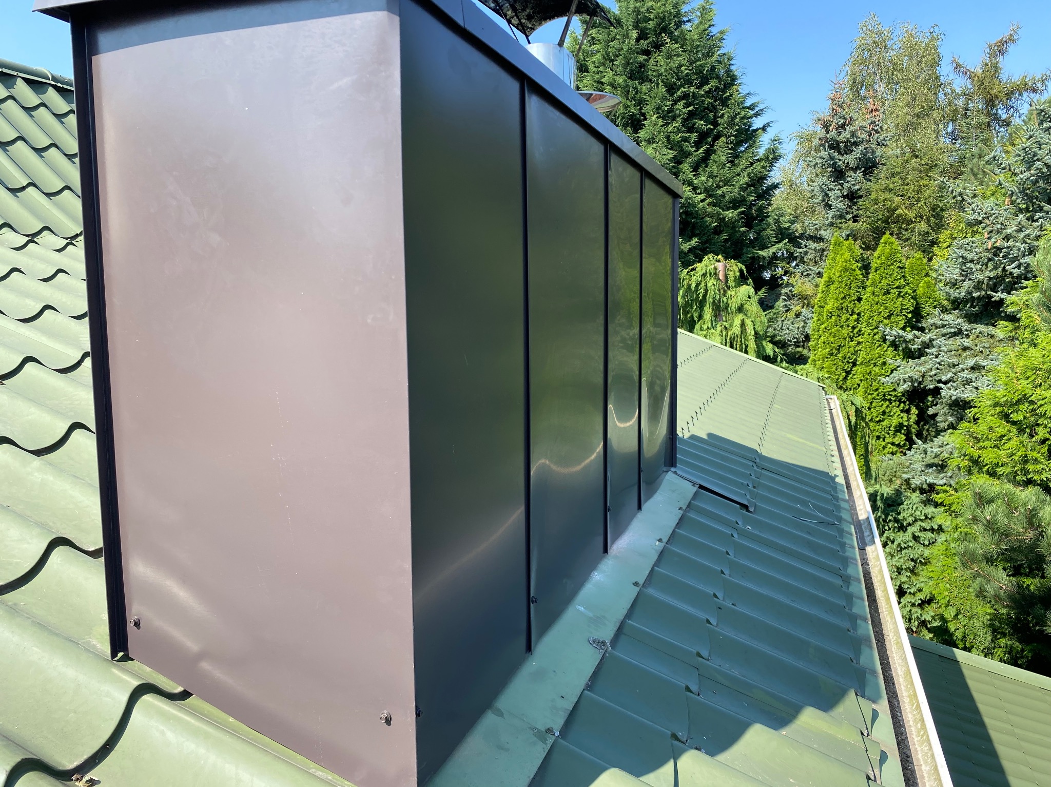 Szczegółowe ujęcie brązowej obróbki komina na zielonym dachu z blachy, na tle zieleni drzew i błękitnego nieba. Widoczne elementy mocujące.