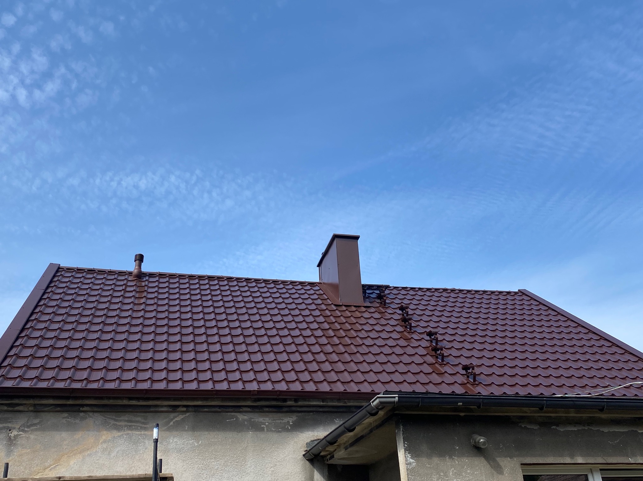 Nowy, brązowy dach z blachodachówki modułowej i kominem na tle błękitnego nieba z delikatnymi chmurami. Widoczne rynny i fragment elewacji budynku.