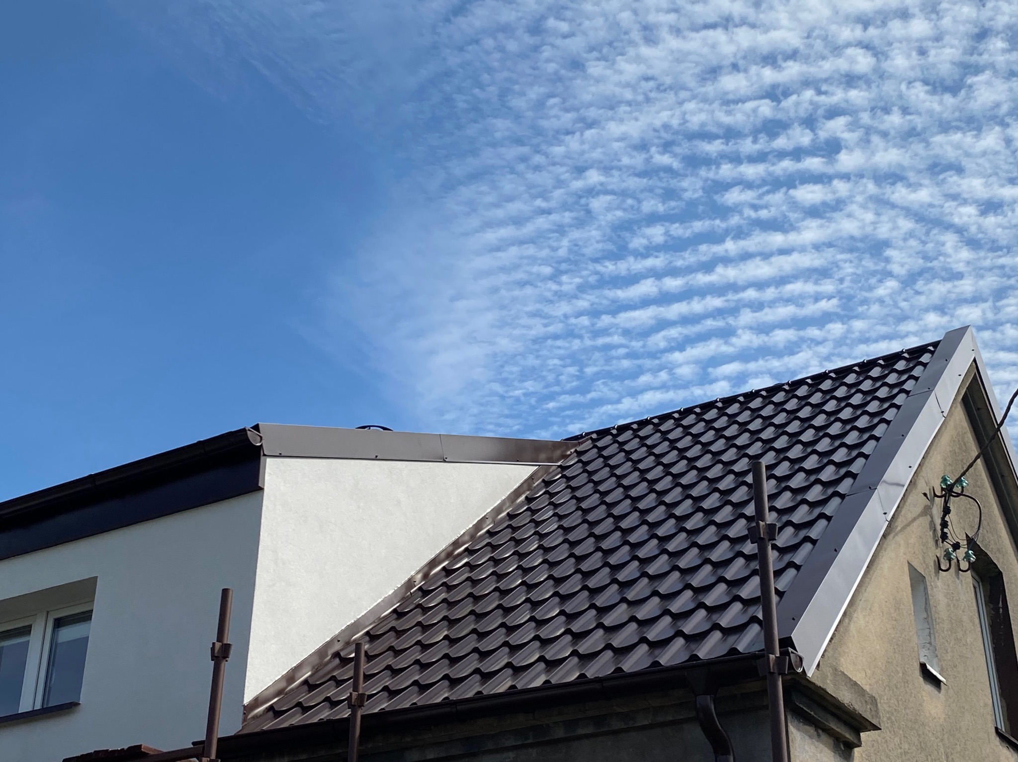 Ciemnobrązowy dach z dachówki ceramicznej na tle błękitnego nieba z chmurami. Widoczne obróbki blacharskie i fragmenty elewacji budynku.