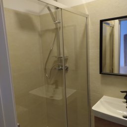 USŁUGI Katarzyna Wasiłowska - Nowoczesna kabina prysznicowa ze szklanymi drzwiami przesuwnymi i chromowaną armaturą. Beżowe płytki ścienne i umywalka z szafką w odcieniach brązu.