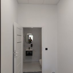 USŁUGI Katarzyna Wasiłowska - Białe wnętrze z otwartymi drzwiami do łazienki po remoncie. Widoczne umywalka, lustro, płytki imitujące marmur i odkurzacz. Na ścianie włącznik światła.