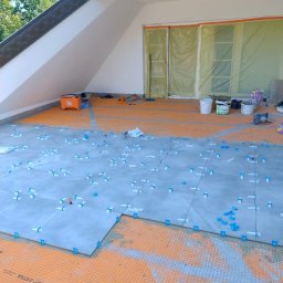 USŁUGI Katarzyna Wasiłowska - Układanie szarych płytek z użyciem niebieskich klipsów poziomujących na pomarańczowej macie uszczelniającej w jasnym, skośnym pomieszczeniu.