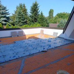 USŁUGI Katarzyna Wasiłowska - Układanie szarych płytek na pomarańczowej macie drenażowej na balkonie z użyciem niebieskich klipsów poziomujących; widok z góry na zewnątrz.