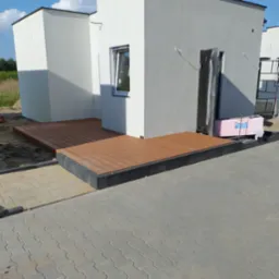 Nowoczesny taras z desek kompozytowych przy białym budynku, z kostką brukową i elementami wykończeniowymi. Widok z zewnątrz, w słoneczny dzień.