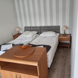 USŁUGI Katarzyna Wasiłowska - Pokój z dwoma łóżkami pojedynczymi, szarą tapetą w pasy i drewnianymi szafkami nocnymi z lampkami. Na stoliku leży okrągła deska. Widok na wyremontowane wnętrze.