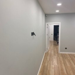 USŁUGI Katarzyna Wasiłowska - Jasne wnętrze po malowaniu: gładkie, szare ściany, drewnopodobna podłoga, białe drzwi i sufit z wbudowanym oświetleniem punktowym. Minimalistyczny design.