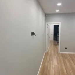 Jasne wnętrze po malowaniu: gładkie, szare ściany, drewnopodobna podłoga, białe drzwi i sufit z wbudowanym oświetleniem punktowym. Minimalistyczny design.